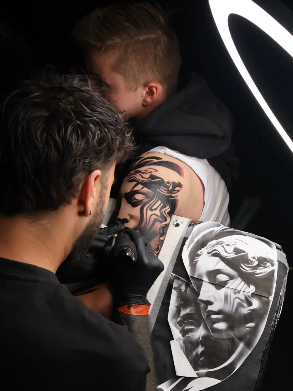 Chesney Foss tatoeëert een klassiek beeldengezicht op de schouder van een klant, met de afgedrukte referentie ernaast